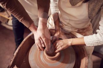 Male Teacher Helping Woman Sitting At Wheel In Pottery Class