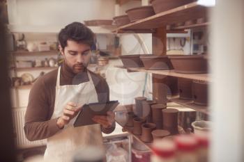 Male Potter In Ceramics Studio Checking Orders Using Digital Tablet