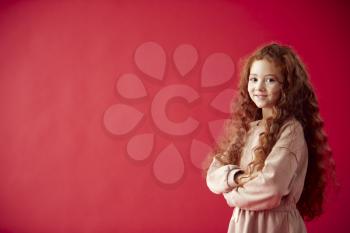 Portrait Of Girl With Long Red Hair Against Red Studio Background Smiling At Camera And Folding Arms