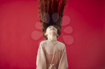 Young Girl With Long Red Hair Tossing It In The Air Against Red Studio Background Smiling At Camera