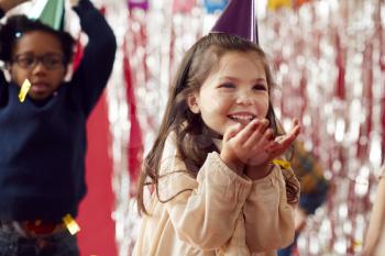 Girl In Party Hat Celebrating At Birthday Party Blowing Handful Of Gold Confetti And Glitter