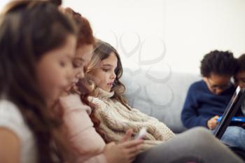 Group Of Children With Friends Sitting On Sofa At Home Playing Together On Digital Devices