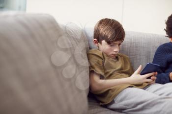 Boy Sitting On Sofa At Home Gaming On Hand Held Devices At Home