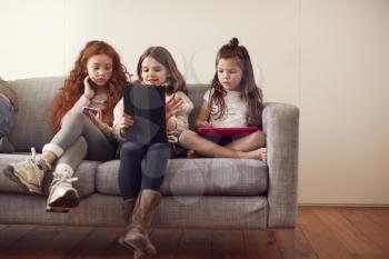Group Of Girls With Friends Sitting On Sofa At Home Playing On Digital Tablet And Mobile Phones
