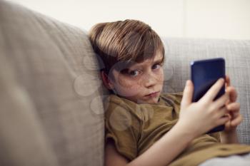 Portrait Of Serious Boy Sitting On Sofa At Home Gaming On Hand Held Devices At Home