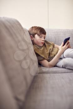 Boy Sitting On Sofa At Home Gaming On Hand Held Devices At Home