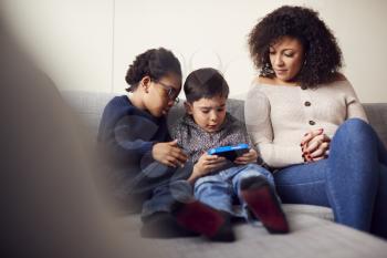 Mother And Sons Sitting On Sofa At Home Playing Computer Game Together On Hand Held Device At Home