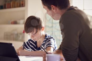 Father Helping Son With Homework Sitting At Kitchen Counter Using Digital Tablet Together