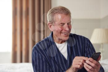 Smiling Senior Man Using Mobile Phone Sitting On Edge Of Bed At Home During Lockdown For Covid-19