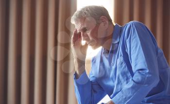 Unhappy And Depressed Senior Man With Head In Hands Sitting On Edge Of Bed At Home