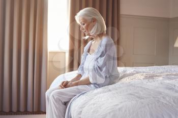 Unhappy And Depressed Senior Woman Sitting On Edge Of Bed At Home
