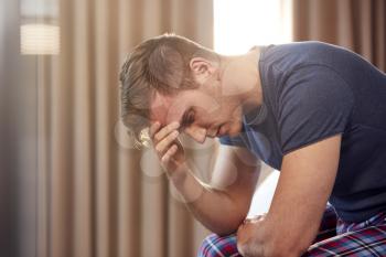 Unhappy And Depressed Man With Head In Hands Sitting On Edge Of Bed At Home