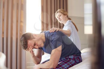 Woman Sitting On End Of Bed At Home Comforting Man Suffering With Mental Health Issues