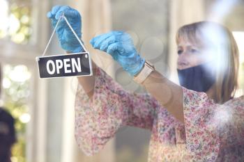 Female Owner Of Coffee Shop Wearing Face Mask Turning Round Open Sign During Health Pandemic