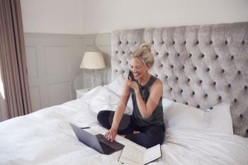 Businesswoman Sitting On Bed With Laptop And Mobile Phone Working From Home During Pandemic Lockdown