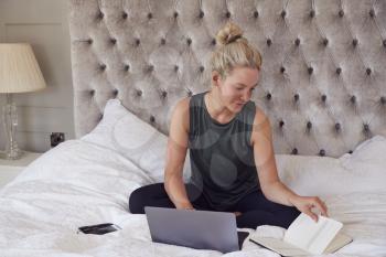 Businesswoman Sitting On Bed With Laptop And Diary Working From Home