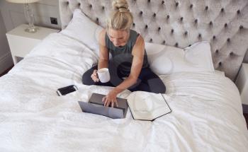 Businesswoman Sitting On Bed With Laptop Working From Home With Hot Drink During Pandemic Lockdown