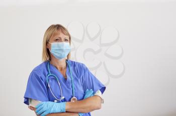 Studio Portrait Of Female Nurse Wearing Scrubs And PPE Face Mask And Gloves Against White Background