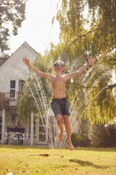 Boy Wearing Swimming Costume Having Fun In Summer Garden Playing In Water From Garden Sprinkler