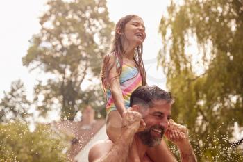 Father Carries Daughter Through Water From Garden Sprinkler Having Fun Wearing Swimming Costumes