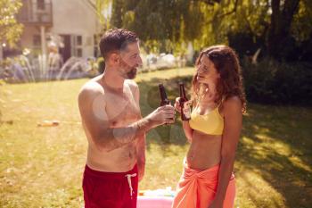 Couple Wearing Swimming Costumes Standing In Paddling Pool In Summer Garden At Home Drinking Beer