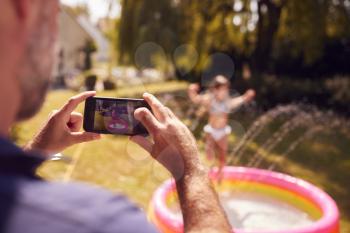 Father Taking Photo Of Daughter Playing In Paddling Pool On Mobile Phone