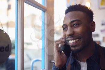 Small Business Owner Standing By Shop Door Making Call On Mobile Phone