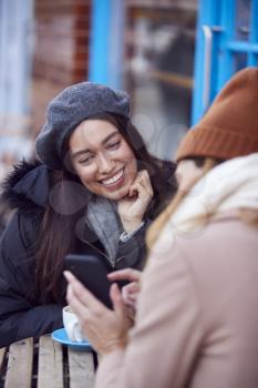 Two Female Friends Meeting Outside Coffee Shop On City High Street Looking At Mobile Phone