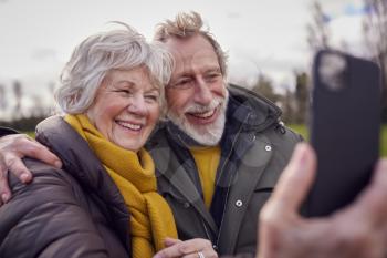 Loving Senior Couple Posing For Selfie On Mobile Phone On Autumn Or Winter Walk Through Park