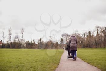 Rear View Of Senior Woman Pushing Senior Man In Wheelchair Outdoors In Fall Or Winter Park