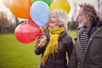 Loving Senior Couple Holding Balloons Enjoying Autumn Or Winter Walk Through Park Together