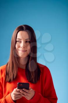 Studio Portrait Of Smiling Young Woman Looking At Mobile Phone Against Blue Background