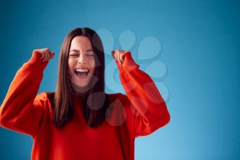 Excited Young Woman Celebrating And Pumping Fists In The Air Against Blue Studio Background