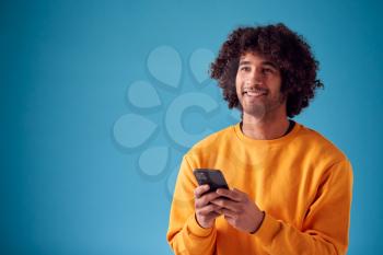 Studio Portrait Of Smiling Young Man Looking At Mobile Phone Against Blue Background
