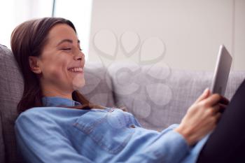 Laughing Young Woman At Home Lying On Sofa Looking At Digital Tablet