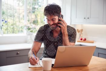 Man In Kitchen With Mobile Phone Working From Home Using Laptop During Health Pandemic