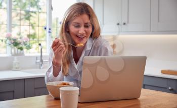 Woman Wearing Pyjamas In Kitchen Watching Laptop Whilst Eating Breakfast
