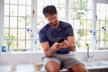 Man Wearing Pyjamas In Kitchen Using Mobile Phone Whilst Eating Breakfast