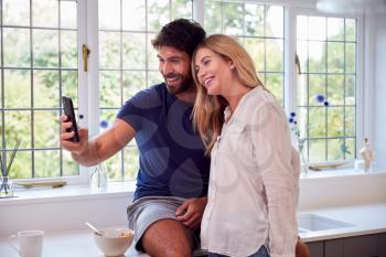 Couple Wearing Pyjamas In Kitchen Using Mobile Phone Whilst Eating Breakfast