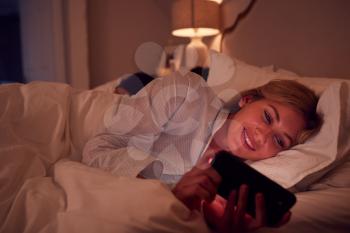 Couple With Woman Lying In Bed At Night Looking At Mobile Phone Screen