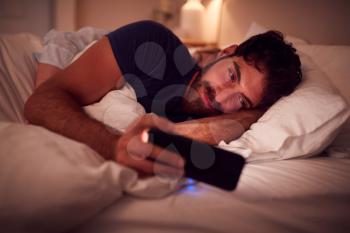Couple With Man Lying In Bed At Night Looking At Mobile Phone Screen