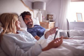 Couple Lying In Bed In Morning Looking At Digital Tablet And Laptop