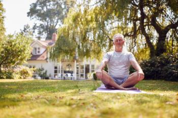 Senior Man At Home In Garden Wearing Fitness Clothing Sitting On Yoga Mat And Meditating