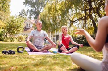 Senior Couple Taking Part In Private Yoga Class With Teacher In Garden At Home