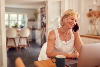 Retired Woman On Phone At Home In Kitchen Using Laptop