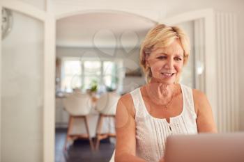 Retired Woman At Home In Kitchen Shopping Online And Managing Domestic Finances Using Laptop
