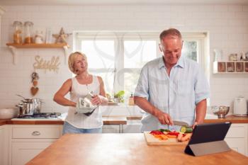 Retired Couple Making Meal In Kitchen With Man Following Recipe On Digital Tablet