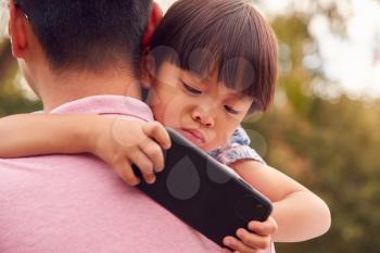 Asian Father Cuddling Son In Garden As Boy Looks Over His Shoulder At Mobile Phone