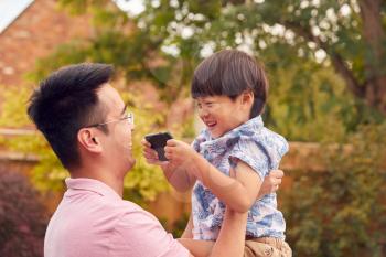 Asian Father Cuddling Son In Garden As Boy Plays With Mobile Phone