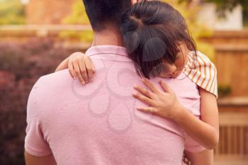 Loving Asian Father Cuddling Daughter In Garden As Girl Looks Over His Shoulder
